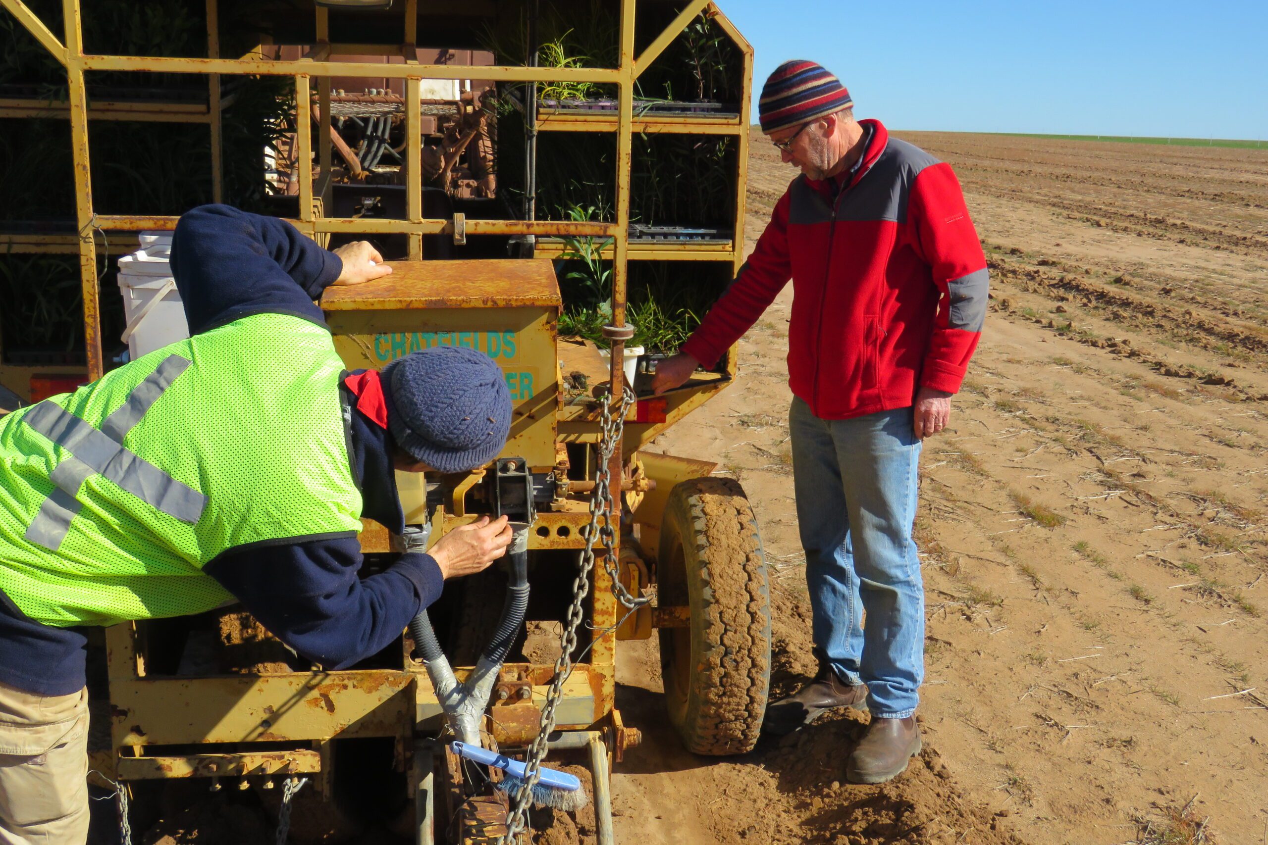 Woman wearing a high-vis vest and beanie with her back to the camera and man wearing red jacket standing at a direct seeding trailer.