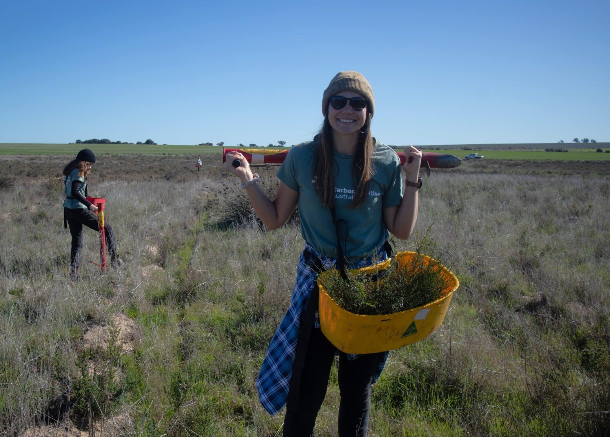Woman holding a bucket of seedlings with another woman planting trees in the background