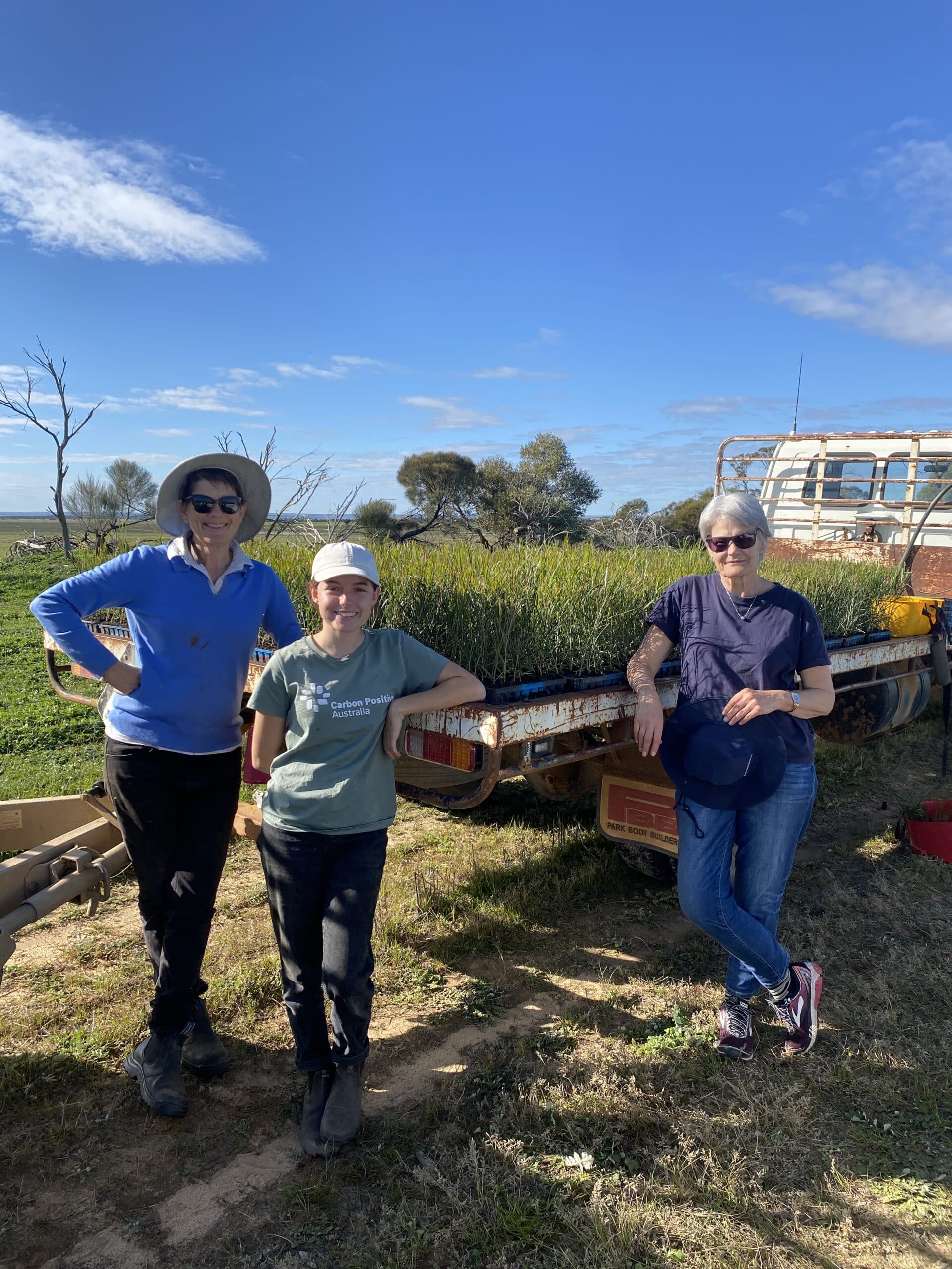 Three women standing and smiling next to a ute tray full of seedlings.