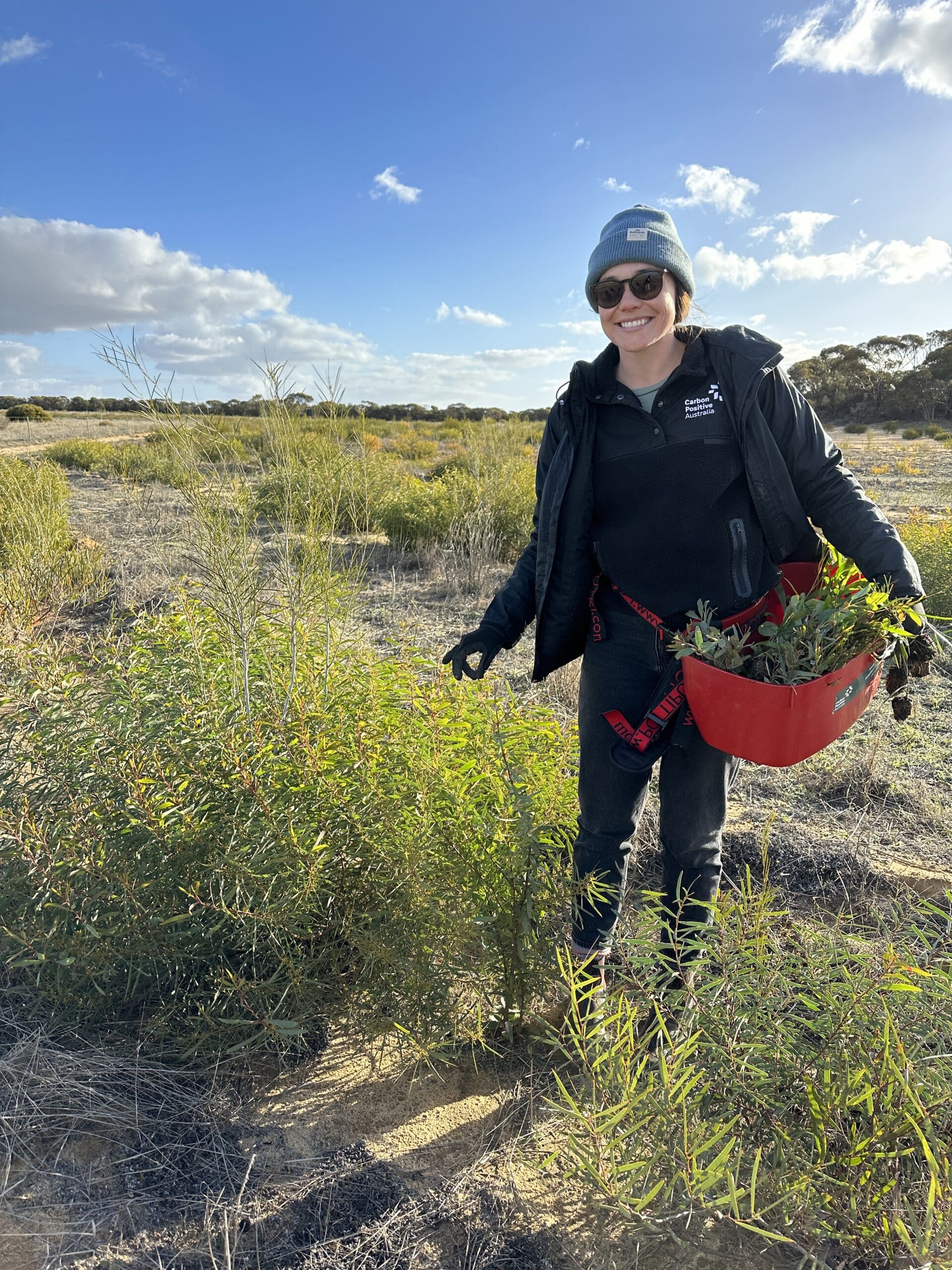 Woman standing smiling next to a shrub with a bucket full of seedlings.