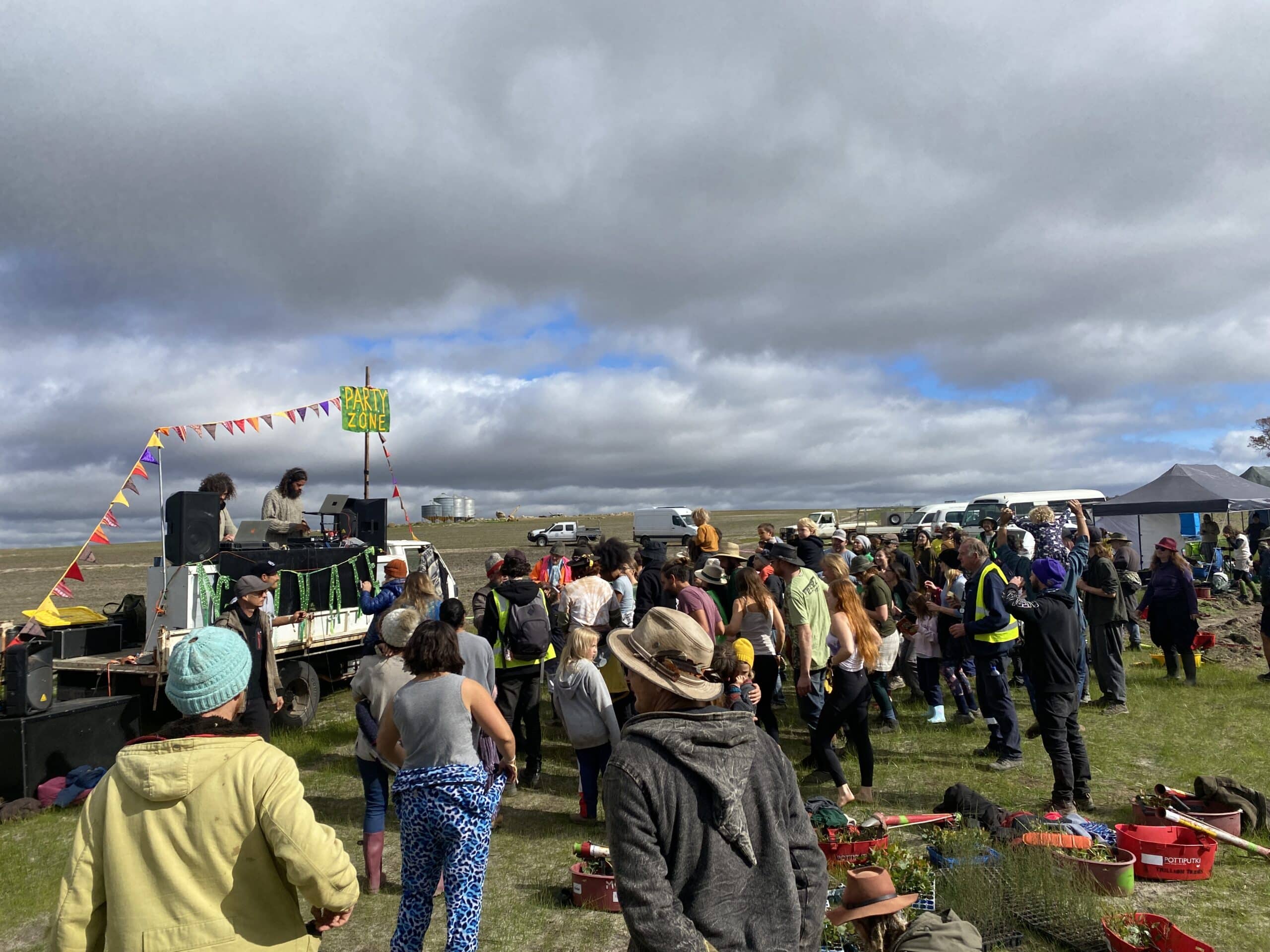 People gathered around in an open paddock dancing to two people playing music from the back of a car.