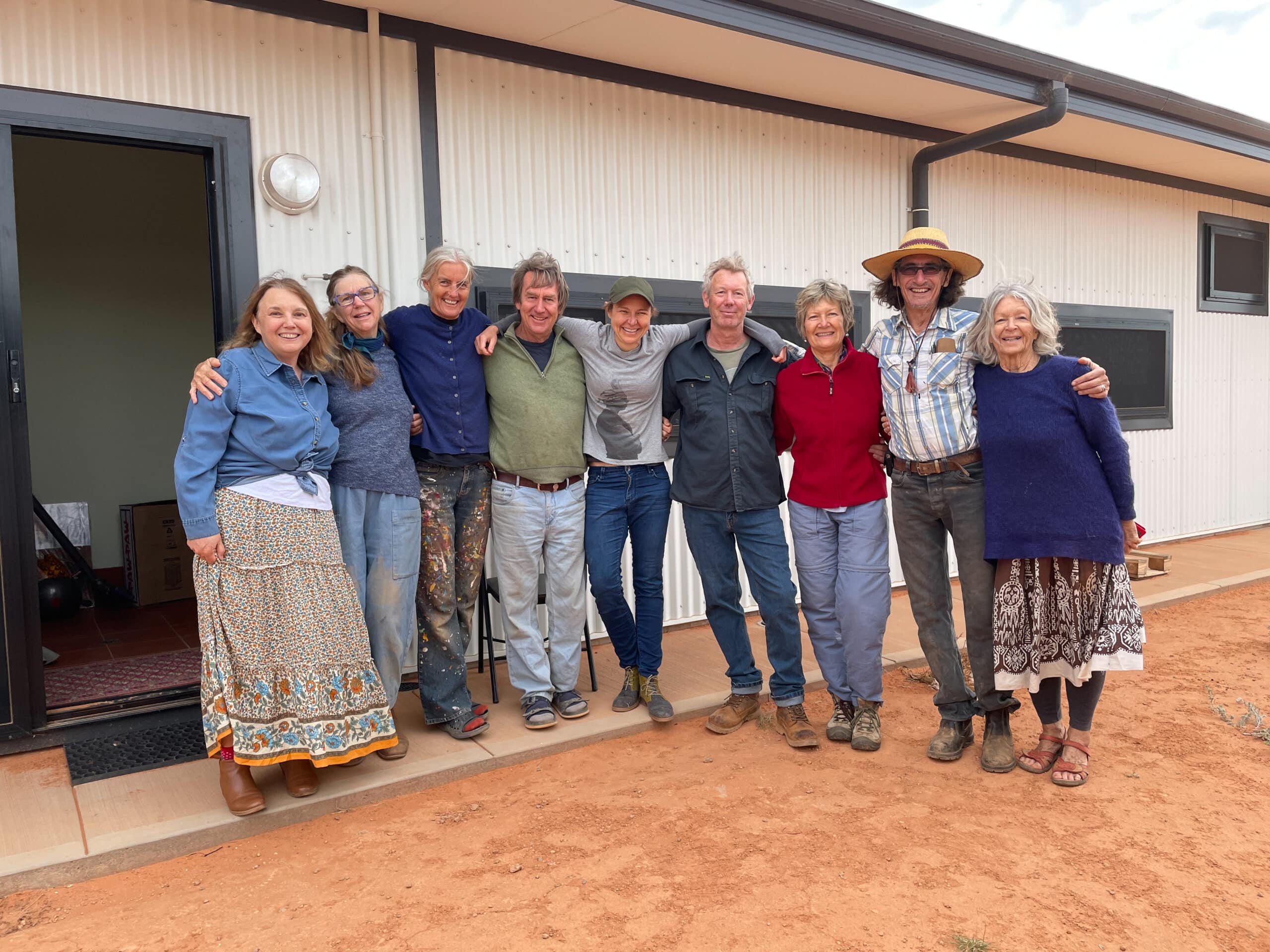 9 people standing and smiling for a photo in from of a building