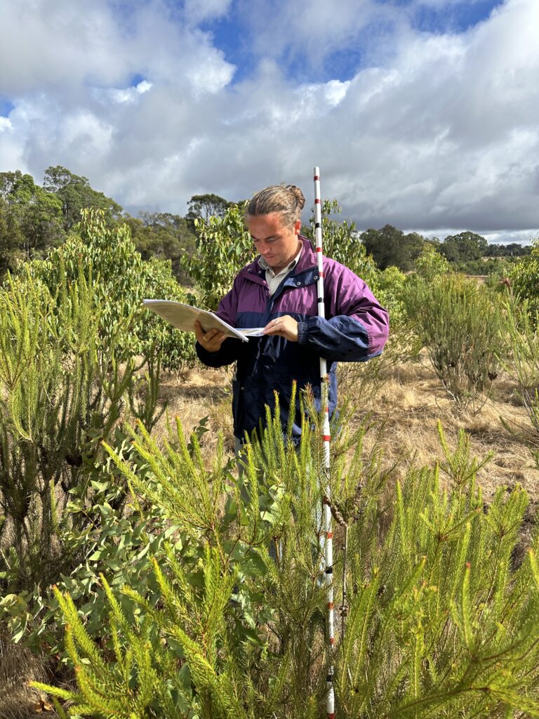 2503_215 Tootanellup Photo 13.JPG Man standing behind a Melaleuca ('Paperbark') tree reading genus toolkit and holding monitoring stick